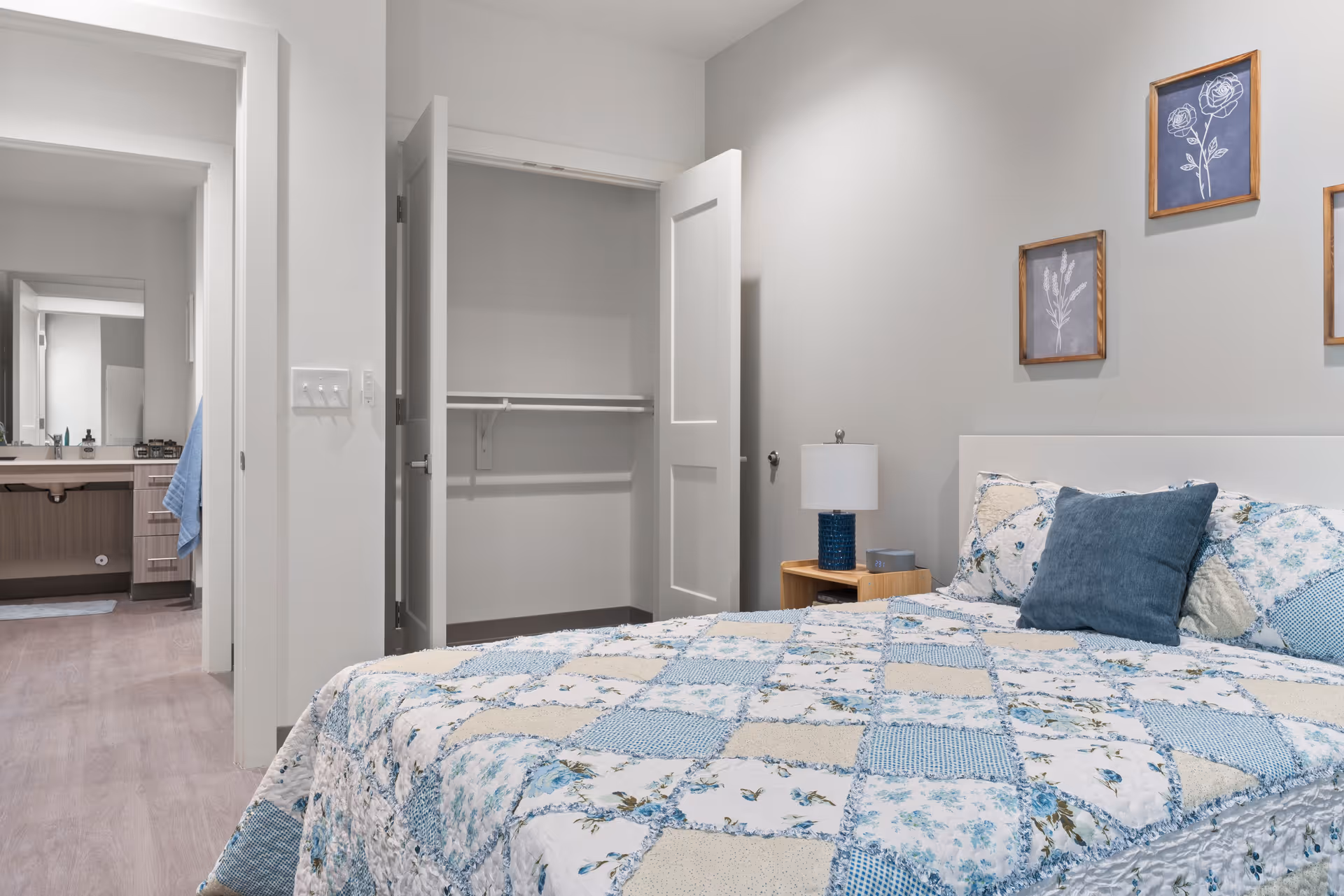A bedroom with a bed covered in a blue and beige patchwork quilt, a blue pillow, and a wooden nightstand with a lamp. The wall behind the bed has three framed floral artworks. To the left, there is an open closet with white doors and an adjacent bathroom with a sink and mirror visible.