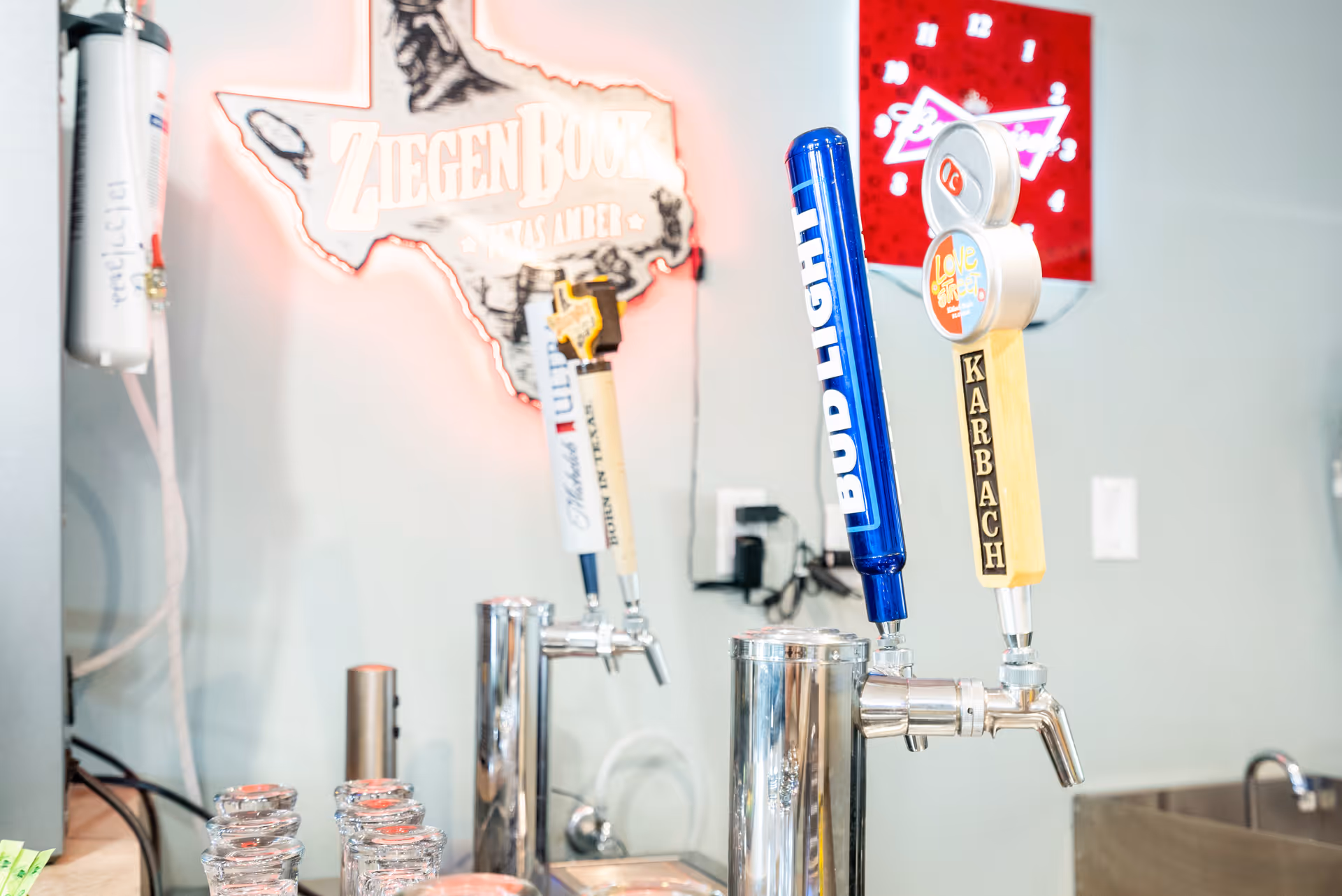 Close-up view of a bar area with several beer taps including Bud Light and Karbach, with a neon sign shaped like Texas in the background that reads ZiegenBock Texas Amber.