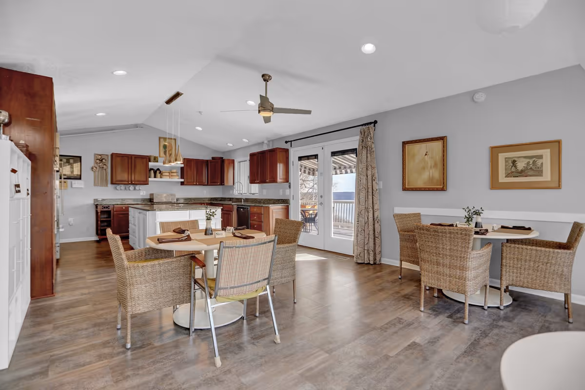 A bright and spacious dining area with two round tables, each surrounded by wicker chairs. The room features a modern kitchen with dark wood cabinets, granite countertops, and stainless steel appliances. French doors with curtains lead to an outdoor patio area. The walls are decorated with framed artwork, and the ceiling has recessed lighting and a ceiling fan.