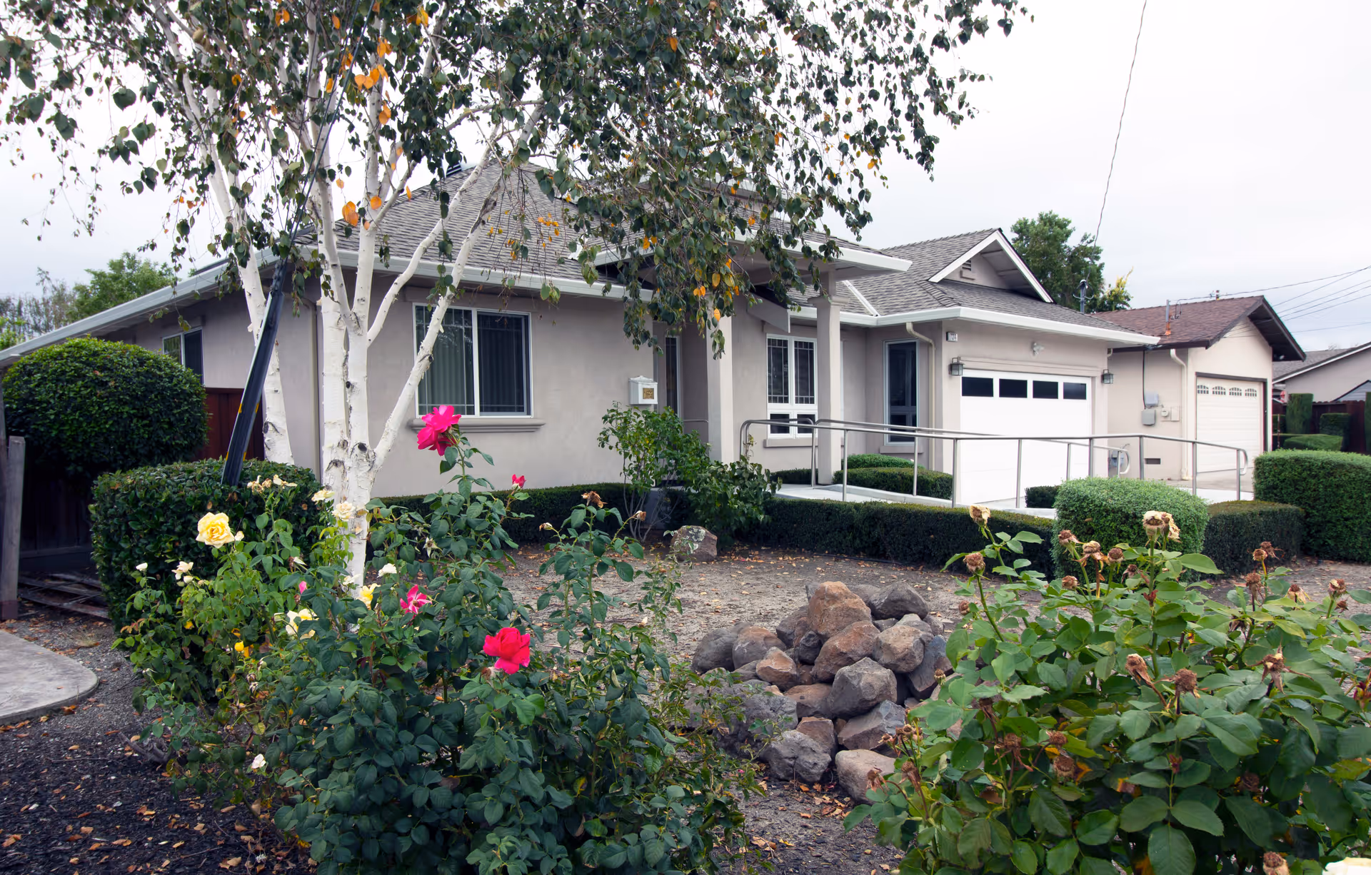Exterior view of a single-story residential building with a ramp leading to the entrance, surrounded by bushes, flowering plants, and a small rock garden in the front yard.