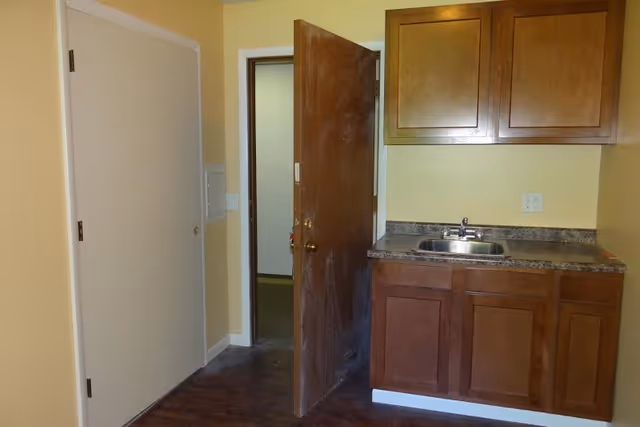 Small kitchenette with wooden cabinets, a countertop sink, an open wooden door, and a sliding closet door.