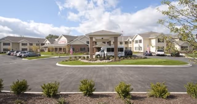 Front exterior view of Amber Park Senior Living facility with a covered entrance, multiple connected buildings, parked cars, and a landscaped area with small bushes and trees under a partly cloudy sky.
