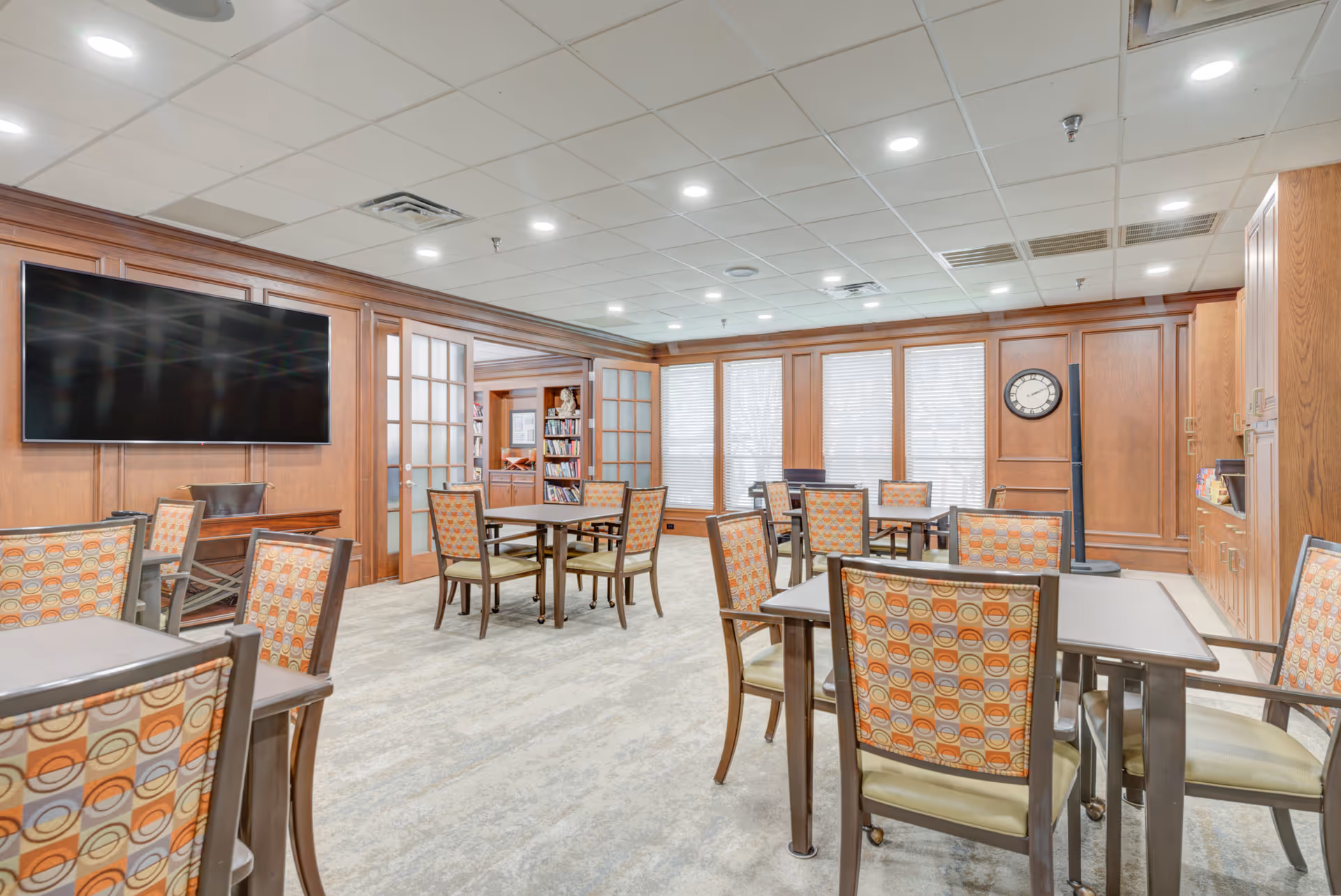 A well-lit common room with multiple tables and chairs featuring patterned upholstery. The room has wood-paneled walls, a large flat-screen TV mounted on one wall, and several windows with blinds. There is a clock on the wall and built-in cabinetry along one side.