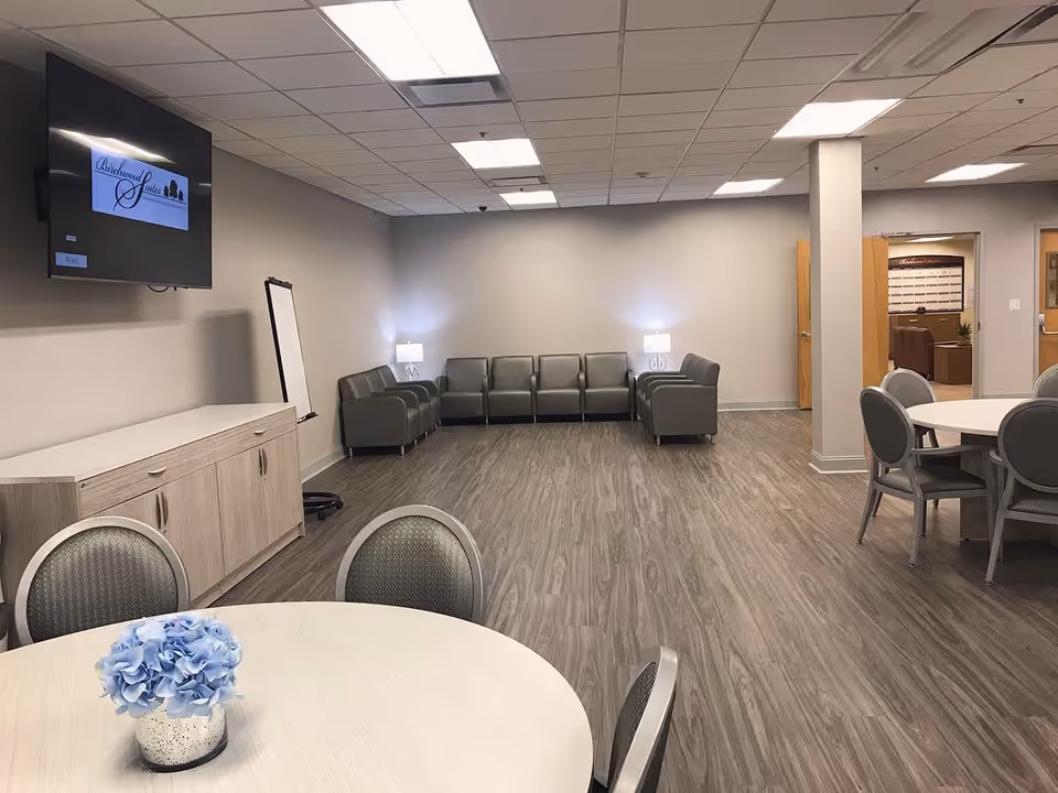 A spacious common area in Birchwood Suites featuring a round table with chairs and a small vase of blue flowers in the foreground, a row of gray armchairs along the back wall with two lamps providing soft lighting, a wall-mounted TV displaying the Birchwood Suites logo, and light wood flooring under a ceiling with recessed lighting.