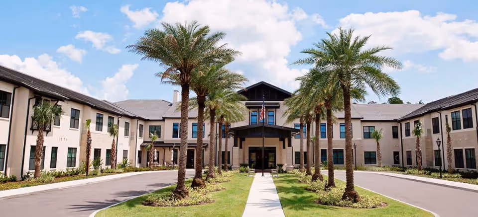 Front exterior of a two-story assisted living building with a driveway and palm trees under a blue sky.