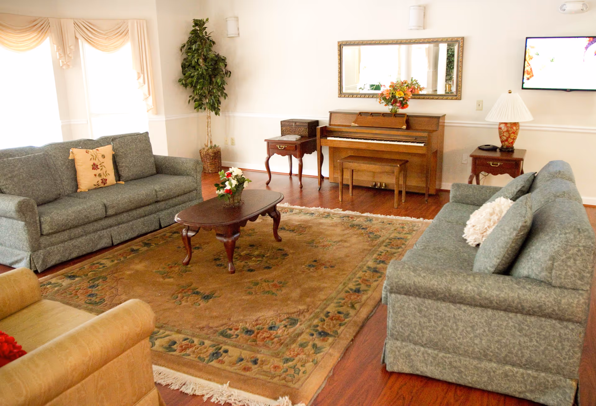 A bright living room with two green sofas facing a wooden coffee table on a large patterned rug, a piano and mirror against the far wall, and side tables with lamps and plants.