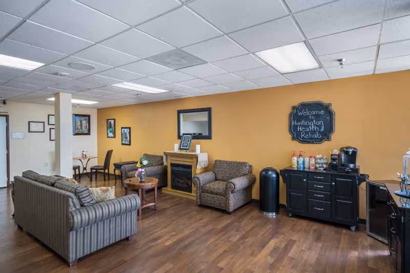 A cozy living room area in Huntington Health and Rehabilitation Center with a striped sofa, an armchair, a small round wooden table with flowers, a fireplace with a mirror above it, and a black cabinet with beverages and a coffee maker. The walls are painted yellow and decorated with framed pictures and a chalkboard sign that reads 'Welcome to Huntington Health & Rehab'.