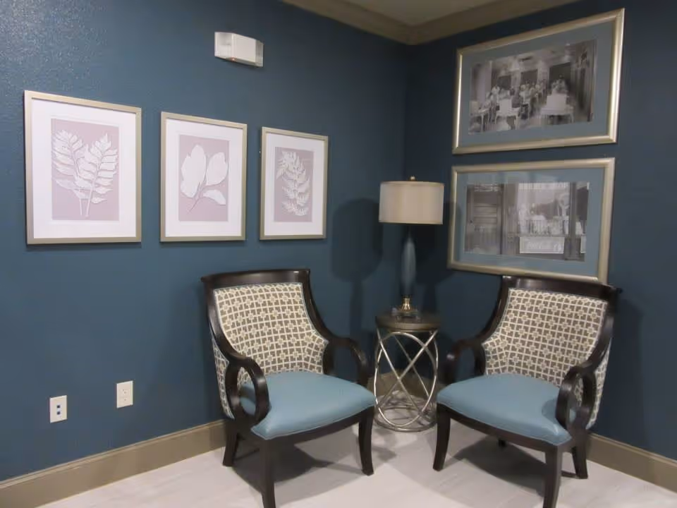 A cozy corner with two patterned armchairs with blue cushions separated by a small round side table with a lamp. The walls are painted dark blue and decorated with five framed pictures, three with botanical prints and two with black and white photographs.