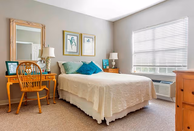 A bright bedroom with a neatly made bed covered in a white bedspread and decorated with teal and blue pillows. On the left side of the bed is a wooden vanity table with a large ornate mirror, a table lamp, and framed photos. A wooden chair is placed in front of the vanity. On the right side of the bed is a wooden nightstand with a white lamp and decorative items. A large window with white blinds lets in natural light, and there is an air conditioning unit below the window. The walls are painted light gray and the floor is carpeted.