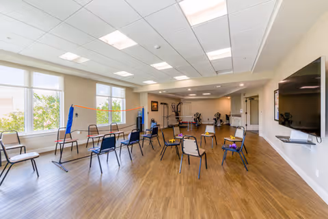 Large multipurpose activity room with spaced chairs in a circle, a low net, exercise equipment at the far end, a wall-mounted TV and large windows letting in daylight.