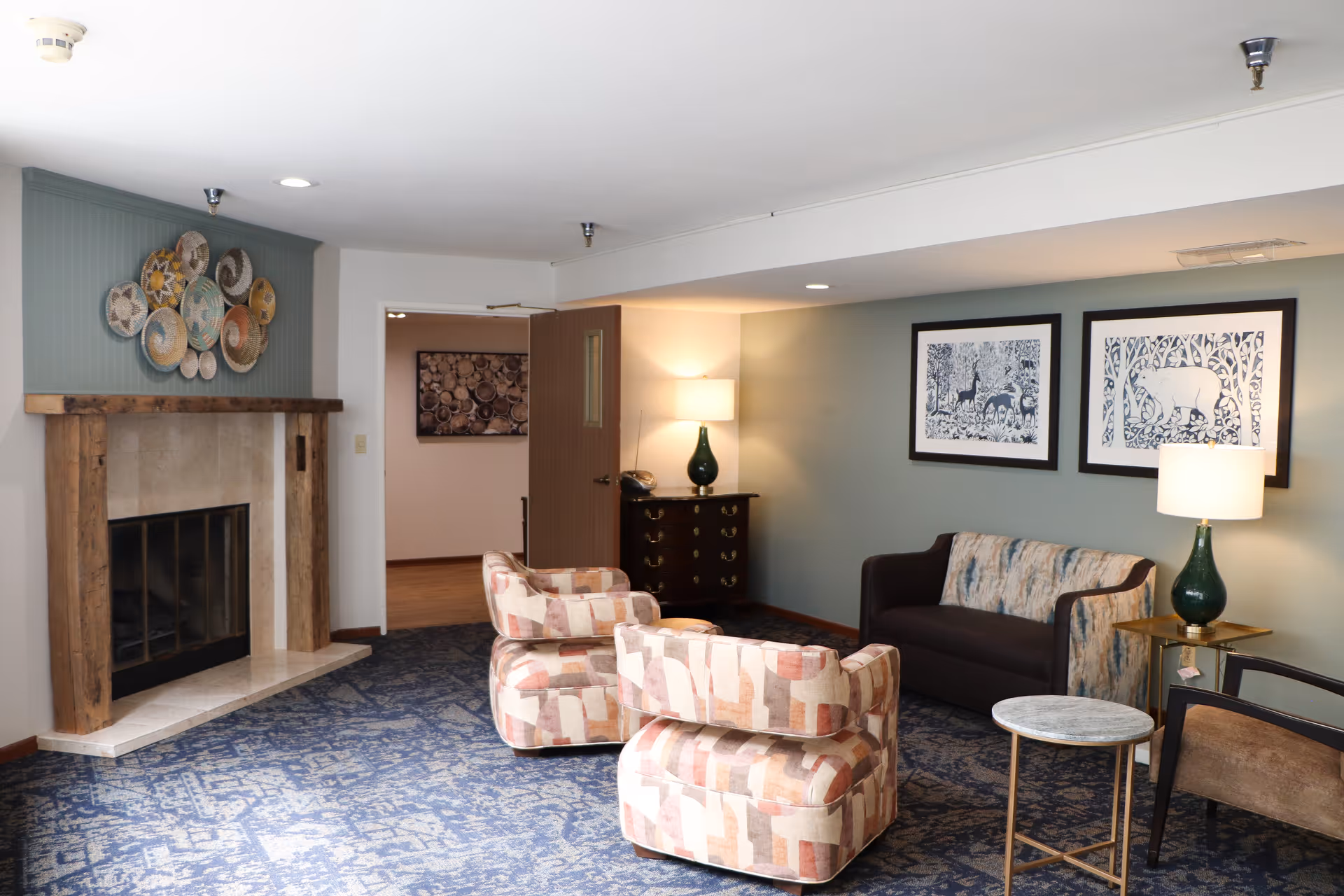 A cozy living room area with a fireplace on the left side featuring a wooden mantel and decorative plates above it. The room has a patterned blue carpet, two patterned armchairs, a dark brown sofa with a patterned cushion, a small round marble-top table, and two green table lamps on side tables. Two framed artworks depicting animals in a forest hang on the wall behind the sofa. A wooden door is open, revealing another room with a wall decoration.