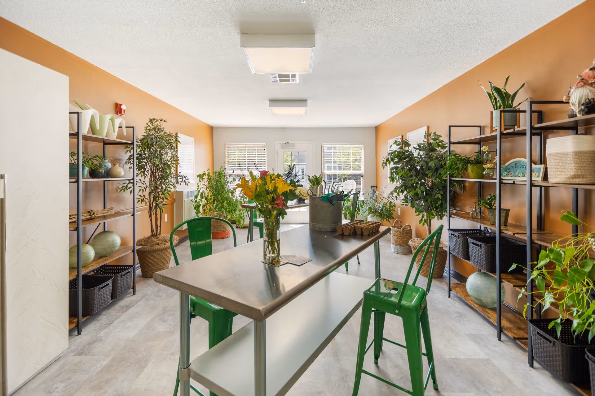 A bright and airy room with orange walls featuring a long metal table with green metal chairs around it. The table has a vase with yellow flowers and gardening supplies. The room is decorated with numerous green plants on black metal shelves and in pots on the floor. Two windows and a glass door at the far end let in natural light.