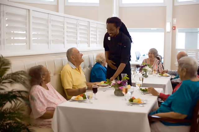 A group of elderly people seated at a long dining table in a bright room, being served by a smiling staff member. The table is set with plates of food, glasses of wine, and floral centerpieces.