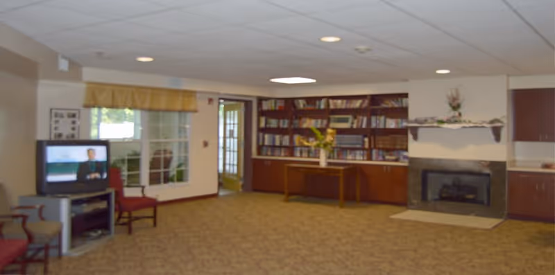 Carpeted communal lounge with chairs, a television, bookshelves, a table with flowers, and a fireplace.