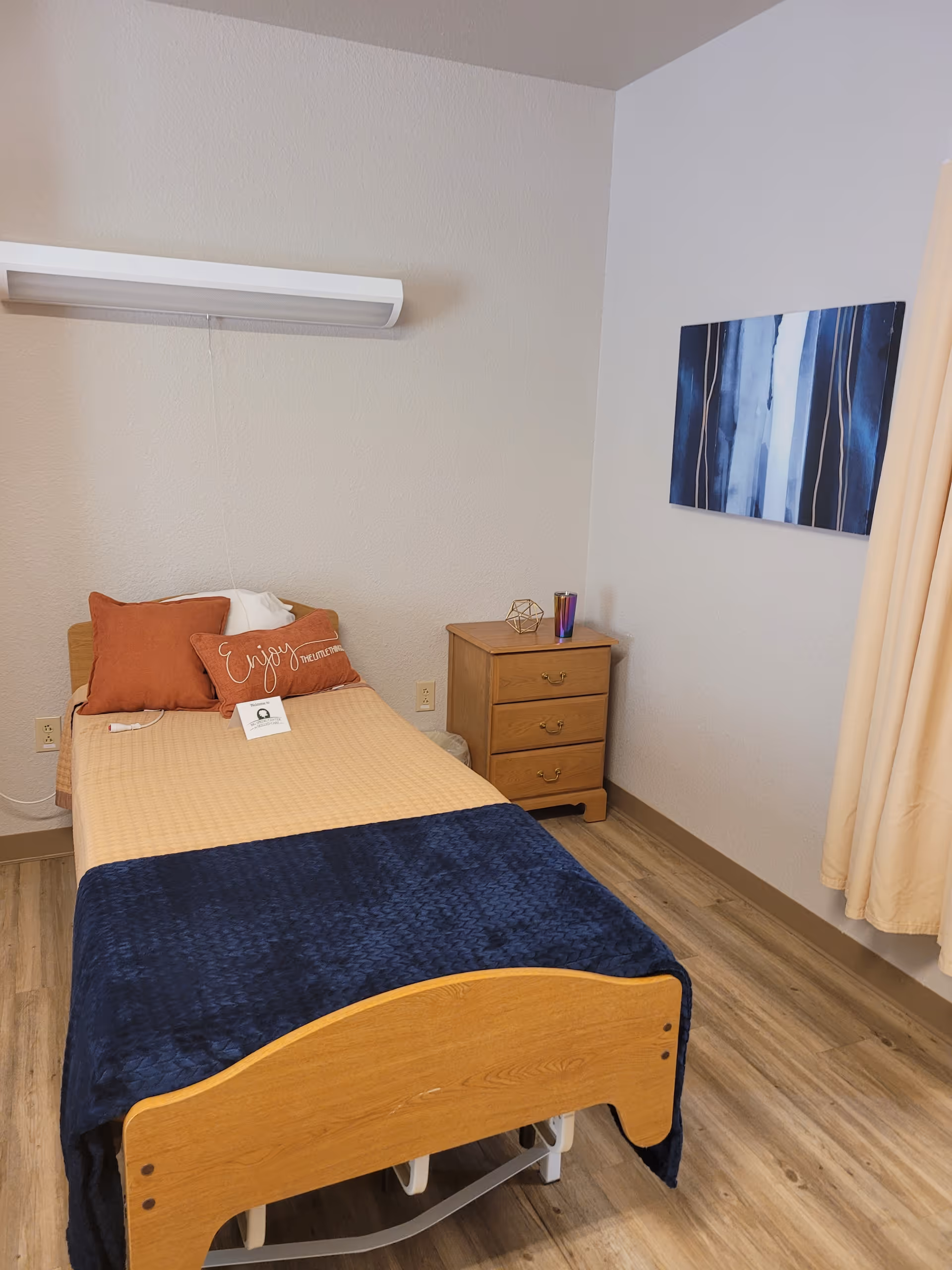 Small resident bedroom with a single bed, navy blanket, orange pillows, a wooden nightstand and wall art on the wall.
