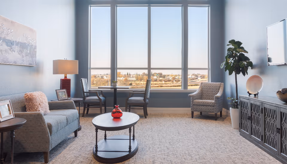 A bright and cozy living room with a large window offering a view of the outdoors. The room features a gray sofa with a pink cushion, a round wooden coffee table with a red vase, two chairs at a small table by the window, a patterned armchair, a floor lamp, a potted plant, and a TV mounted on the wall above a decorative cabinet.