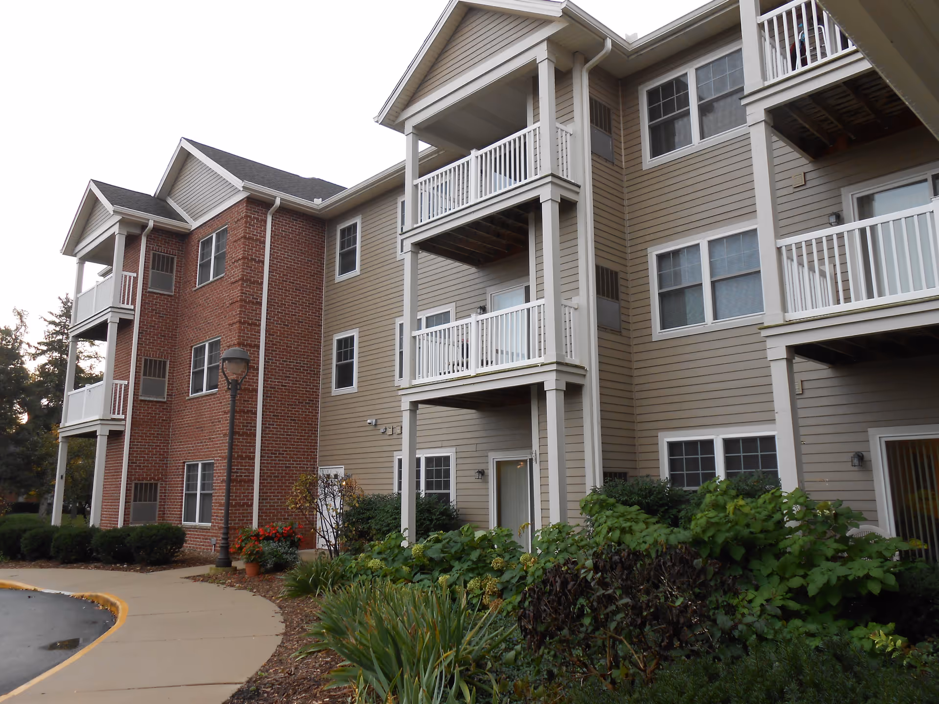 Exterior view of a multi-story residential building with beige siding and red brick accents. The building features balconies with white railings, several windows, and a landscaped area with bushes and plants along a curved sidewalk.