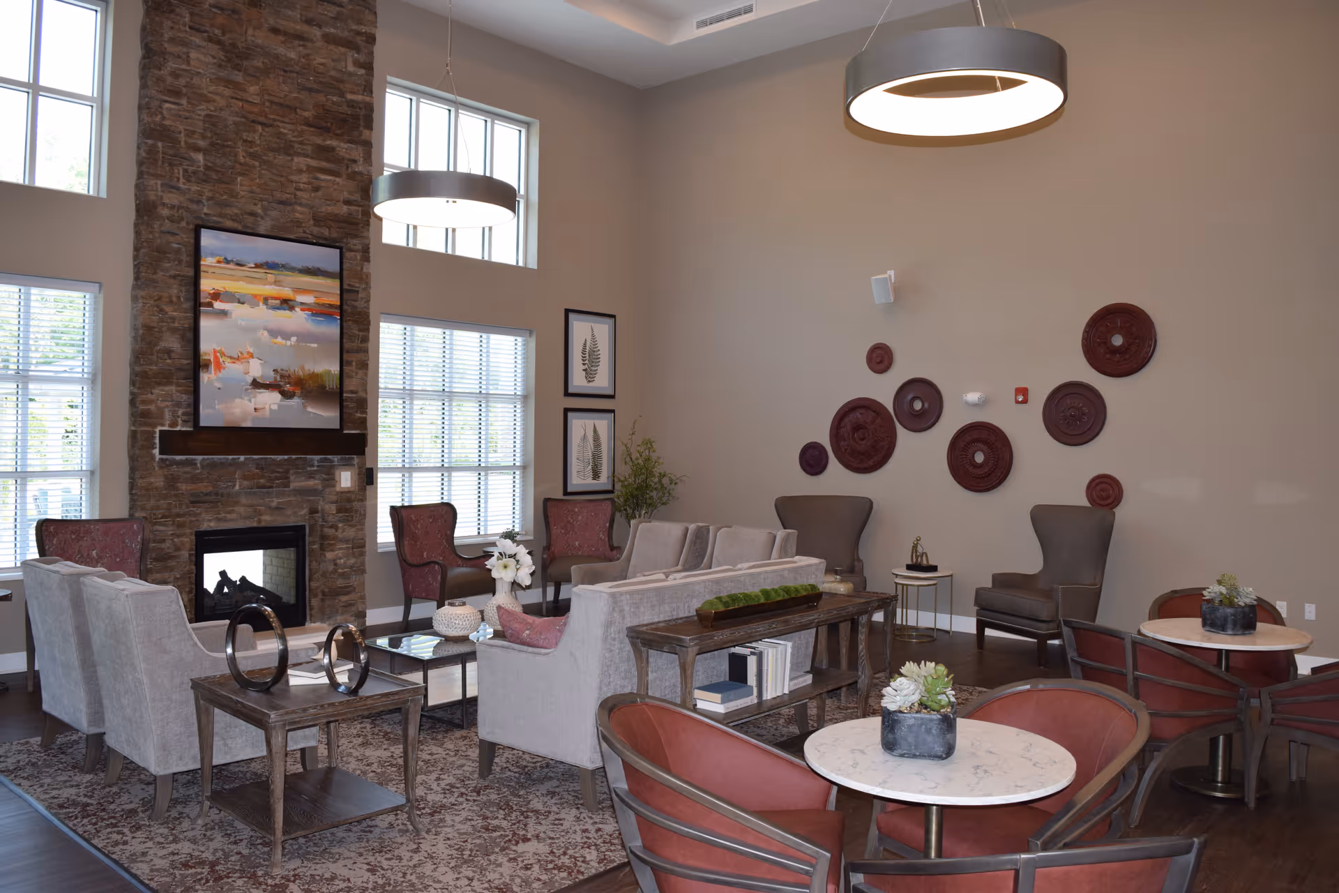 A spacious and well-lit living room area with large windows and a stone fireplace. The room features multiple seating options including beige sofas, red armchairs, and gray wingback chairs. There are decorative wall hangings, framed botanical prints, a patterned rug, and modern circular pendant lights hanging from the ceiling. Small tables with plants and books add to the cozy atmosphere.