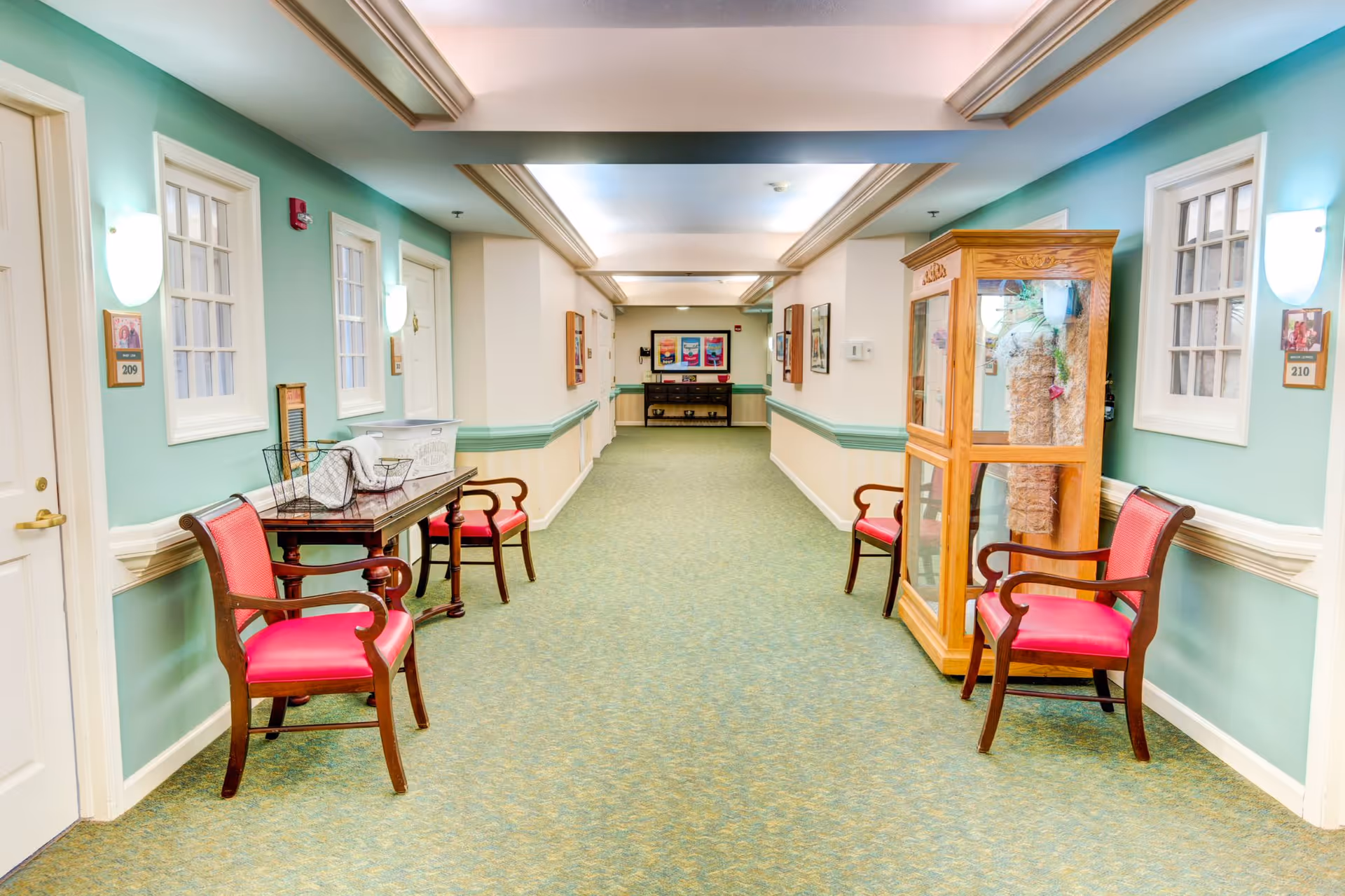 A brightly lit hallway in a senior living facility with teal and cream walls, green carpet, and wooden chairs with red cushions placed along the sides. There are doors with room numbers 209 and 210, a wooden display cabinet, and framed pictures on the walls.