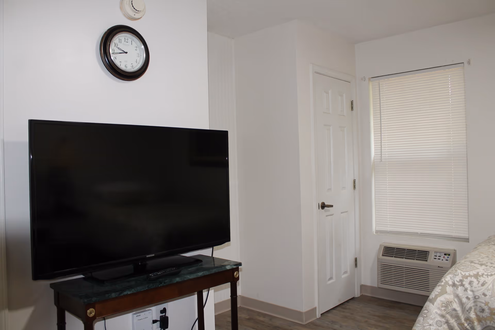 A corner of a bedroom with a flat-screen TV on a wooden table, a wall clock above the TV, a closed white door, a window with closed blinds, and a wall-mounted air conditioning unit below the window. Part of a bed with patterned bedding is visible on the right side.