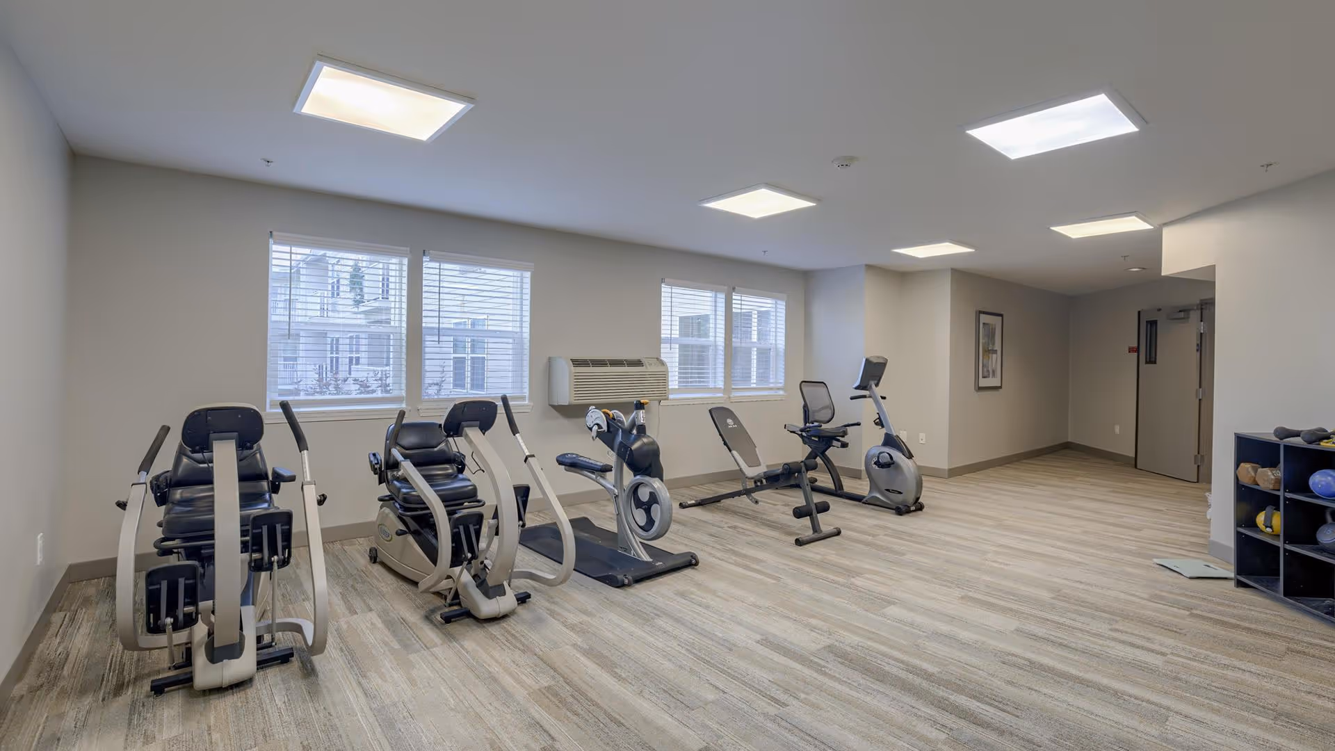 A fitness room with exercise equipment including two recumbent bikes, a rowing machine, a weight bench, and an upright stationary bike. The room has light-colored walls, wood-patterned flooring, three windows with blinds, and ceiling lights. There is a small shelving unit with exercise balls and weights on the right side.