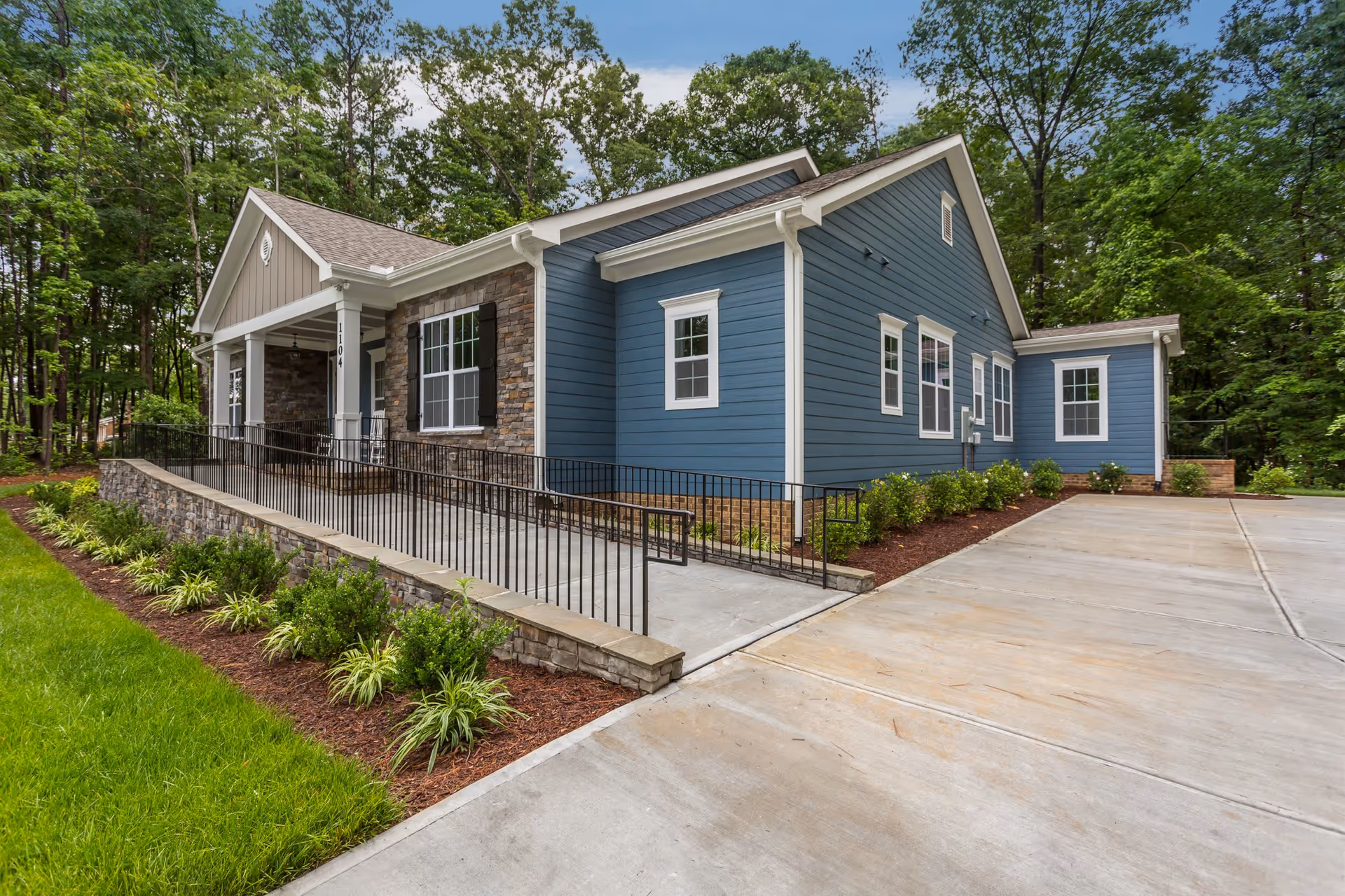 Exterior view of a single-story assisted living facility building with blue siding and stone accents. The building has a covered porch with a ramp and black metal railing, surrounded by landscaped greenery and trees in the background. The driveway and walkway are made of concrete.