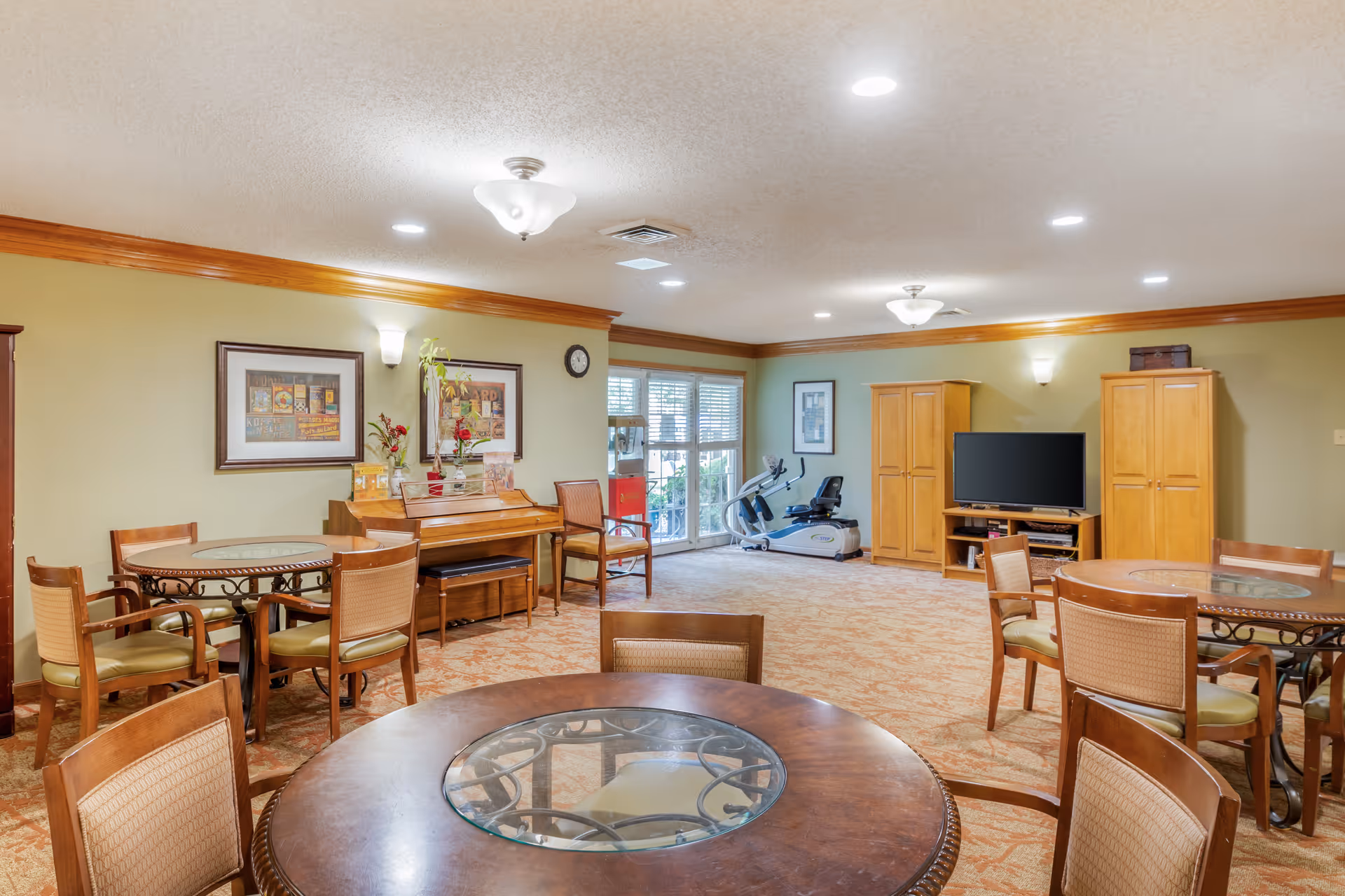 A spacious common room with multiple round tables and chairs, a piano against the wall with framed artwork above it, a television set between two wooden cabinets, and an exercise bike near a window with blinds. The room has beige walls with wooden trim and carpeted flooring.
