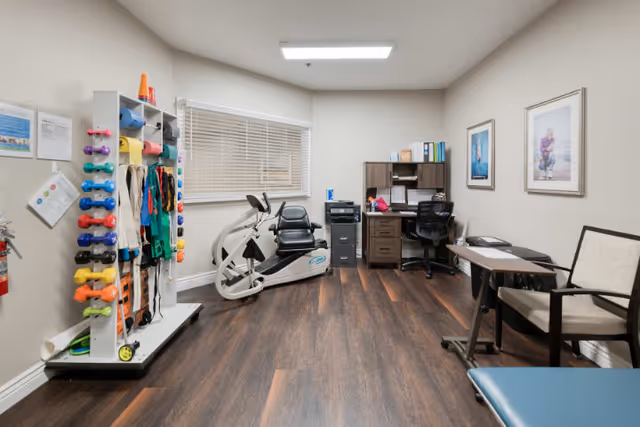 A small fitness and therapy room with exercise equipment including a recumbent bike, a rack of colorful dumbbells and resistance bands, a desk with office supplies, two chairs, and framed pictures on the wall. The room has wood flooring and a window with blinds.