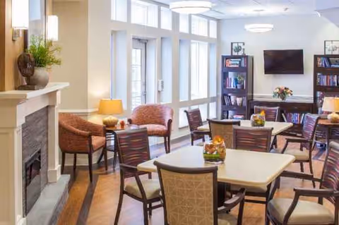 Bright senior living lounge with cream chairs, tables, fireplace, bookshelves, and large windows.