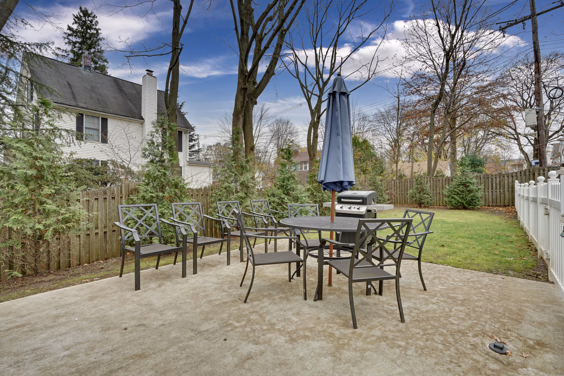 Outdoor patio area with a round metal table, several matching chairs, a closed blue umbrella in the center of the table, and a barbecue grill. The patio is surrounded by a wooden fence, trees, and a grassy yard with a white fence on the right side. A house is visible in the background under a partly cloudy sky.