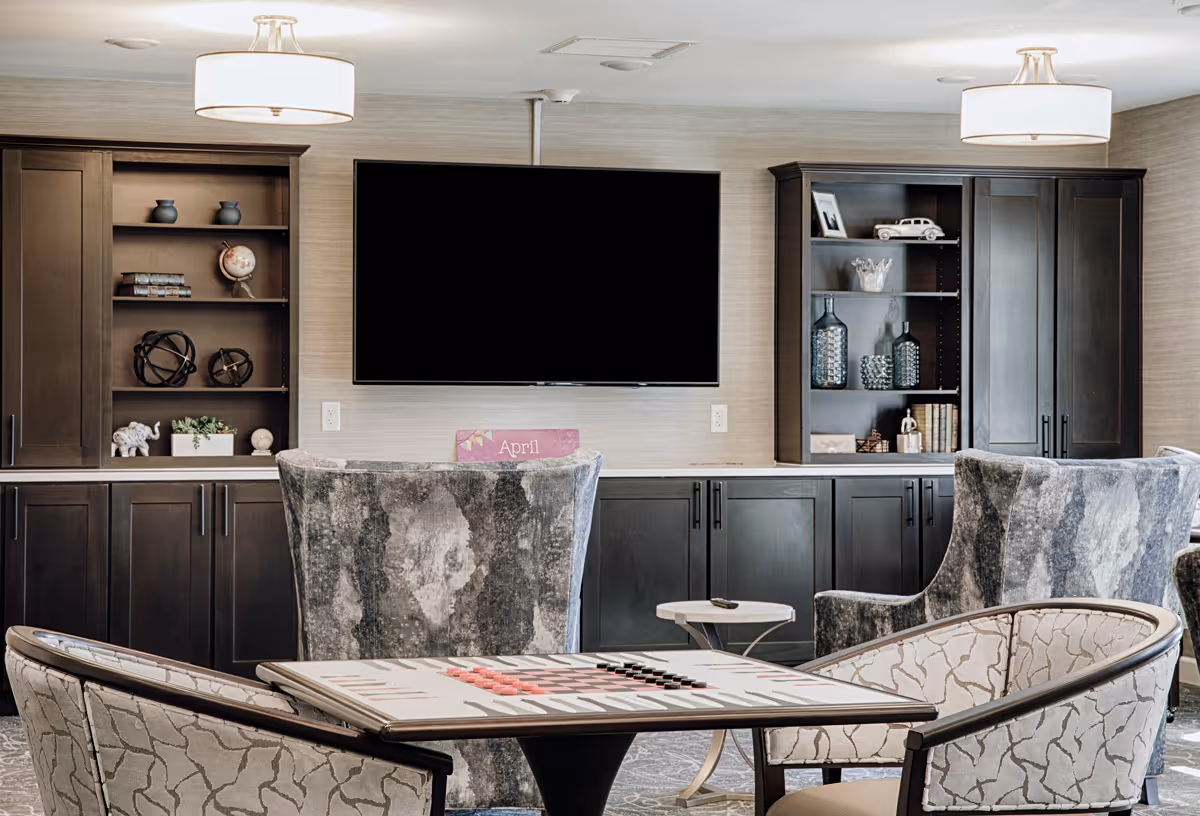 A cozy common area with a large flat-screen TV mounted on the wall, flanked by dark wooden cabinets with shelves displaying decorative items. In front of the TV, there are comfortable upholstered chairs and a table with a backgammon board set up for play. The room is softly lit by ceiling lights.