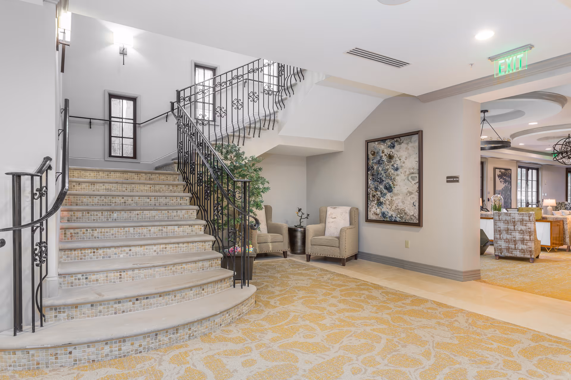 A bright and spacious interior area featuring a curved staircase with decorative tile risers and black wrought iron railing. Two beige armchairs with a small table and a potted plant are positioned near the staircase. A large abstract painting hangs on the wall. The floor is covered with a patterned carpet in shades of yellow and beige. In the background, a seating area with additional chairs and tables is visible. An illuminated green exit sign is mounted on the ceiling.