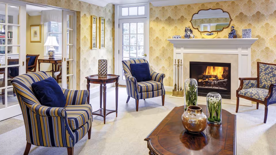 A cozy living room with a lit fireplace, two striped armchairs with dark blue cushions, a wooden coffee table with decorative vases and glass domes containing flowers, a side table with a decorative lantern, and a patterned armchair. The room has yellow patterned wallpaper, a mirror above the fireplace, and French doors leading to another room with a desk and lamp.
