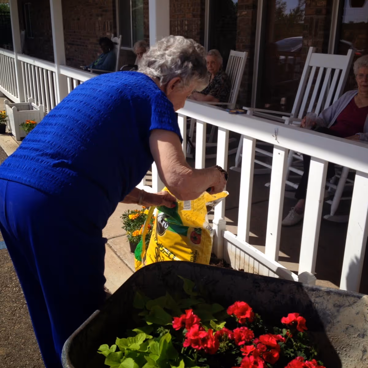 An elderly woman pours potting soil into a planter on a porch while others sit in rocking chairs and red flowers sit in a nearby wheelbarrow.