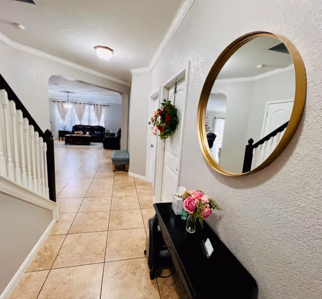 Hallway with tiled floor, a black console table topped with flowers and a round wall mirror, a staircase to the left and a living room visible ahead.