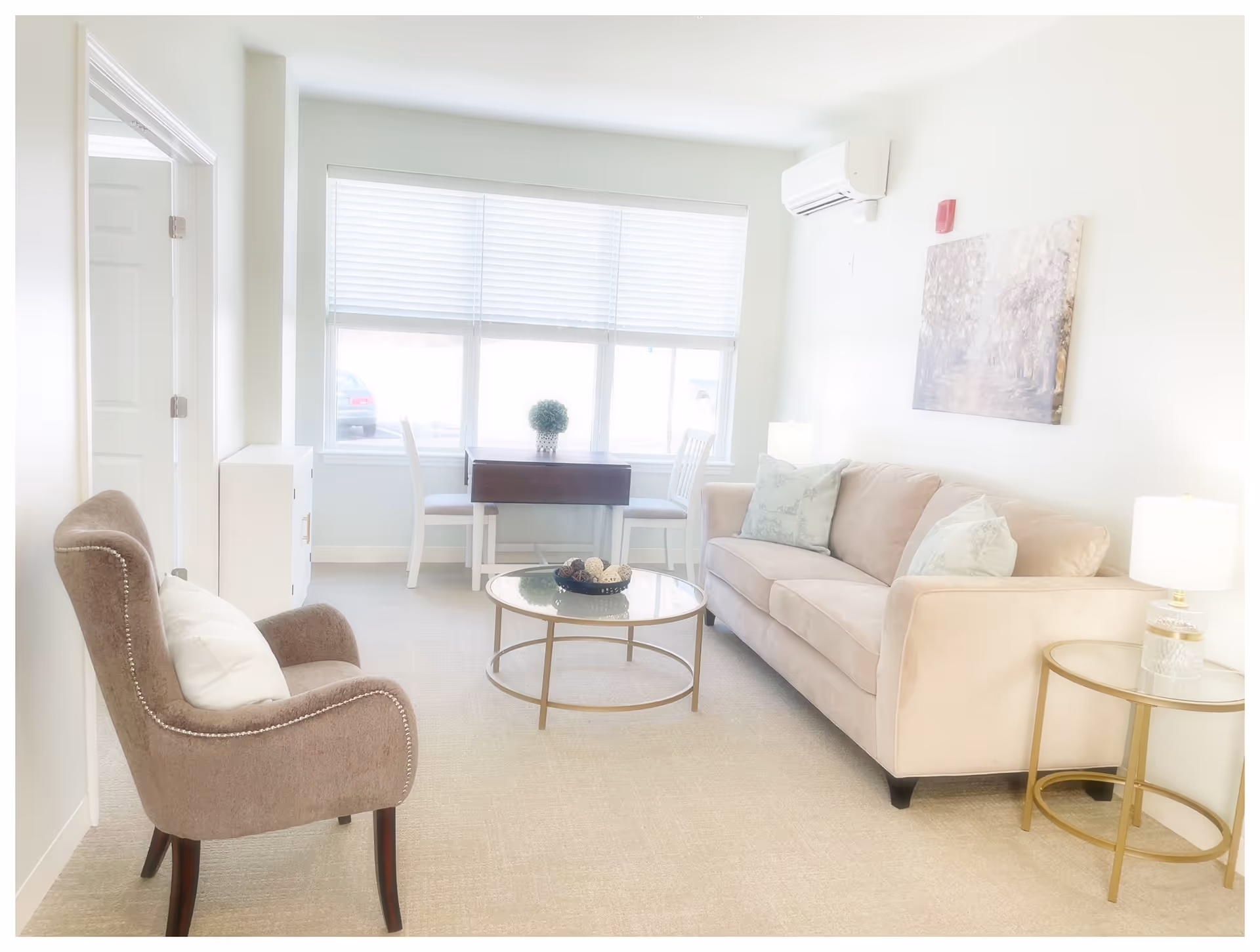 A bright living room with a beige sofa adorned with light-colored pillows, a round glass coffee table with decorative items, a beige upholstered armchair with a white pillow, a small round side table with a lamp, and a small dining table with two white chairs near a large window with blinds.