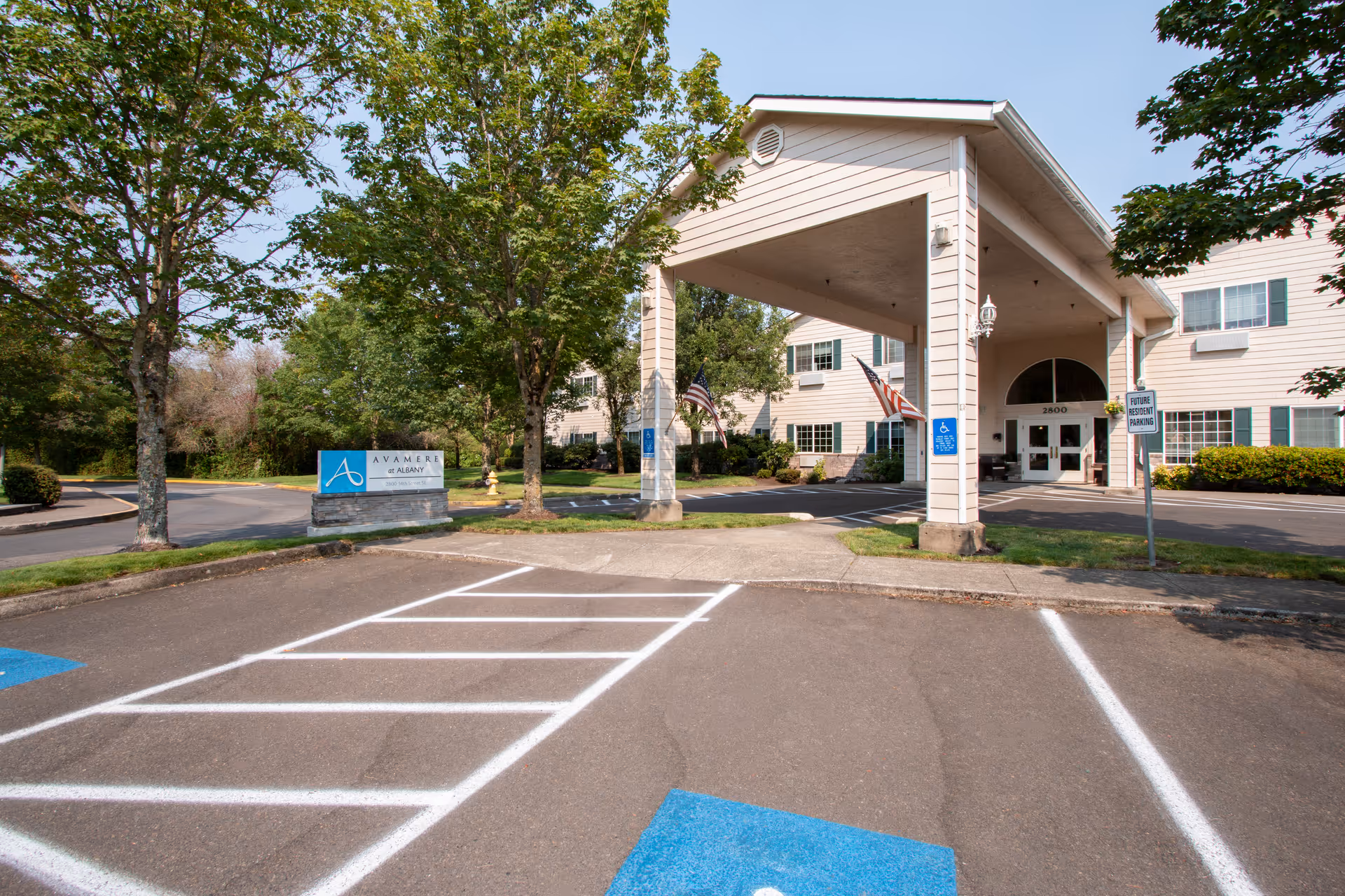Exterior view of a senior living facility entrance with a covered drop-off area, handicap parking spaces, trees, and a sign that reads 'Avamere at Albany'. The building is light-colored with multiple windows and American flags near the entrance.