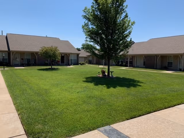 A well-maintained grassy courtyard with a few small trees and a birdbath in the center, surrounded by single-story brick buildings with covered patios and rocking chairs under a clear blue sky.