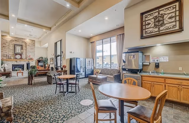 A spacious common area in a senior living facility with patterned carpet and tiled floor. The room features round wooden tables with chairs, a sofa and coffee table near large windows with curtains, vending machines, a kitchenette with a coffee machine and sink, and a fireplace with a brick surround and decorative items on the mantel.