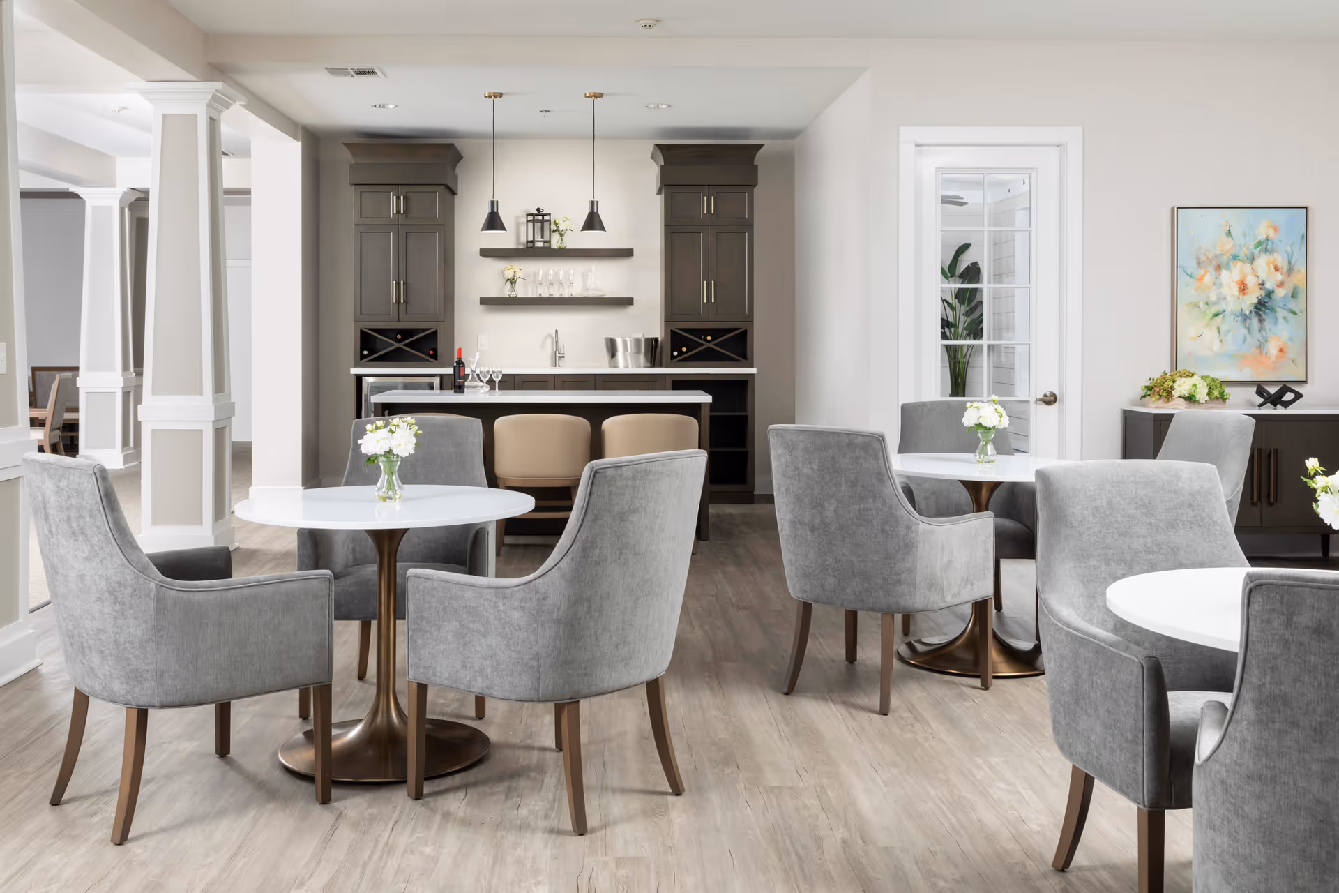 A bright and modern dining area with round white tables and gray upholstered chairs. Each table has a small vase with white flowers. In the background, there is a kitchenette with dark wood cabinets, a countertop with two beige stools, pendant lights, and shelves holding glassware and decorative items. A door with glass panels and a sideboard with a floral painting and plants are also visible.