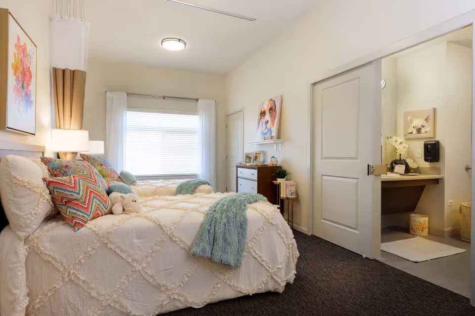 A cozy bedroom with two twin beds covered in white textured bedspreads and colorful pillows. A small stuffed animal is placed on one of the beds. The room has a window with white curtains, a dresser with framed photos, and a door leading to a bathroom with a sink and a wall-mounted soap dispenser. The walls are decorated with colorful animal paintings.
