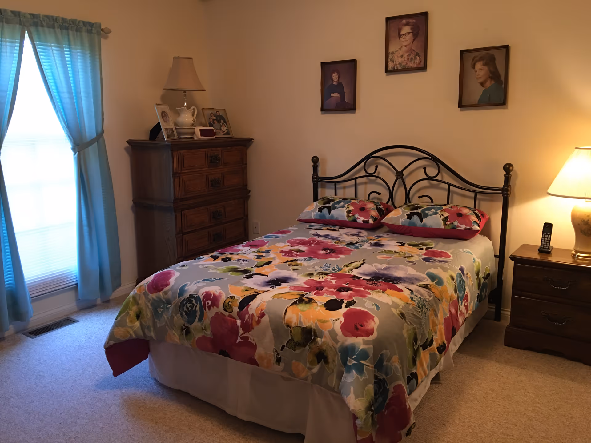 A bedroom with a bed featuring a colorful floral bedspread and two matching pillows. The bed has a black metal headboard. To the right of the bed is a wooden nightstand with a lamp and a cordless phone. On the left side of the bed is a tall wooden dresser with a lamp, a teapot, and framed photos on top. Three framed portraits hang on the wall above the bed. A window with blue curtains is on the left side of the room.