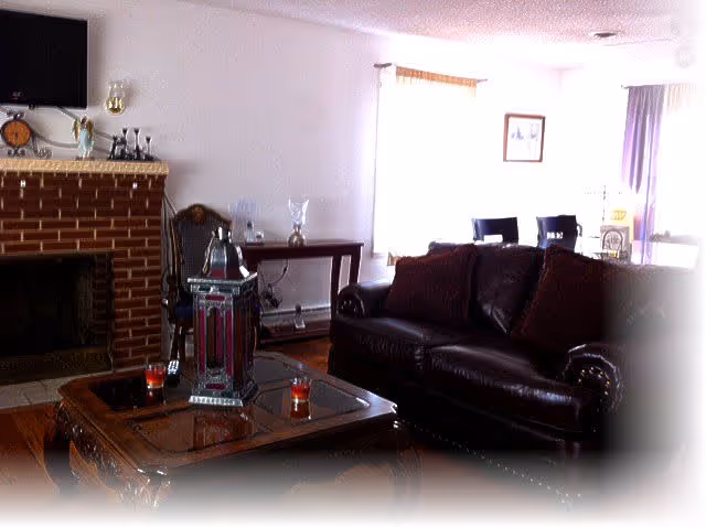 A cozy living room with a dark leather sofa adorned with cushions, a wooden coffee table with decorative items including candles and a lantern, a brick fireplace with a mantel holding various ornaments, a wooden chair, and a dining area with chairs visible in the background near windows with curtains.