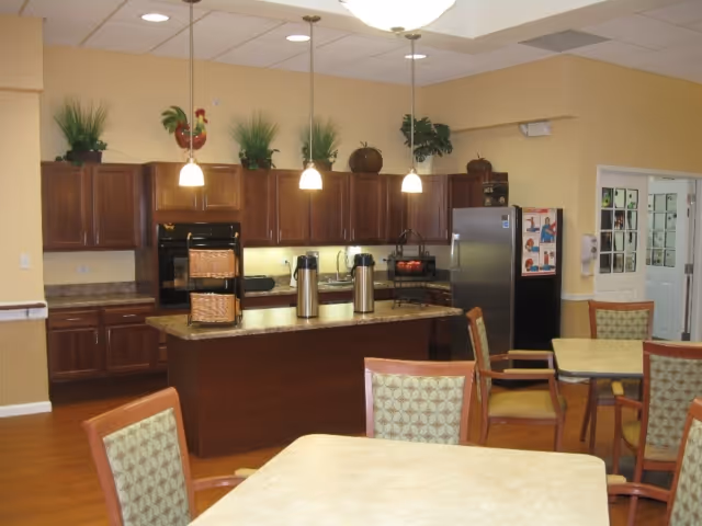 Communal kitchen and dining area with a central island and pendant lights, wooden cabinets, refrigerator, and several dining tables and chairs.
