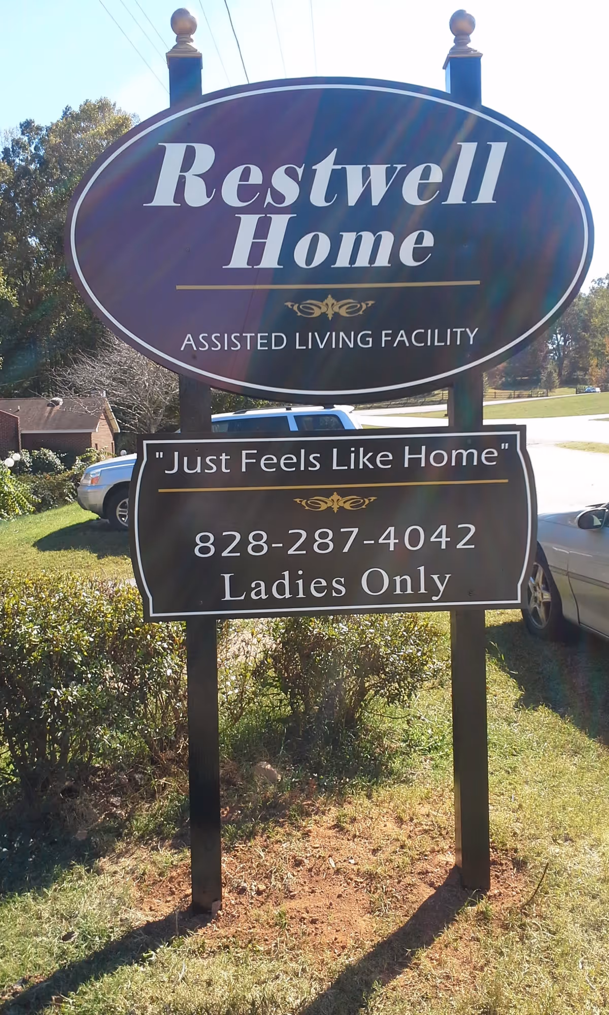 A roadside sign reading "Restwell Home Assisted Living Facility" and "Ladies Only" with a phone number in front of a grassy lawn and parked cars.