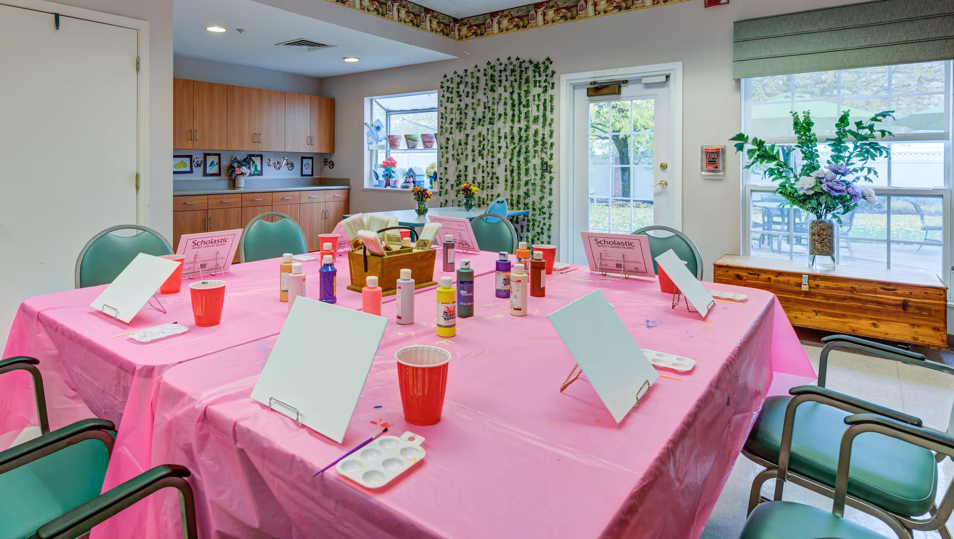 A brightly lit activity room with a large table covered in a pink plastic tablecloth. The table is set up for painting with blank canvases on easels, paint bottles, paint palettes, and red plastic cups. Surrounding the table are green cushioned chairs. In the background, there are wooden cabinets, a window with potted plants, a door leading outside, and a wooden chest with a vase of flowers on top.