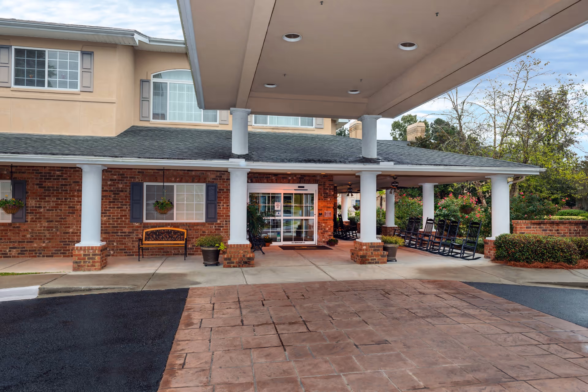 Entrance of The Bridge at Charleston facility showing a covered drop-off area with white columns, brick walls, a bench, potted plants, and several black rocking chairs on the right side under the covered porch.