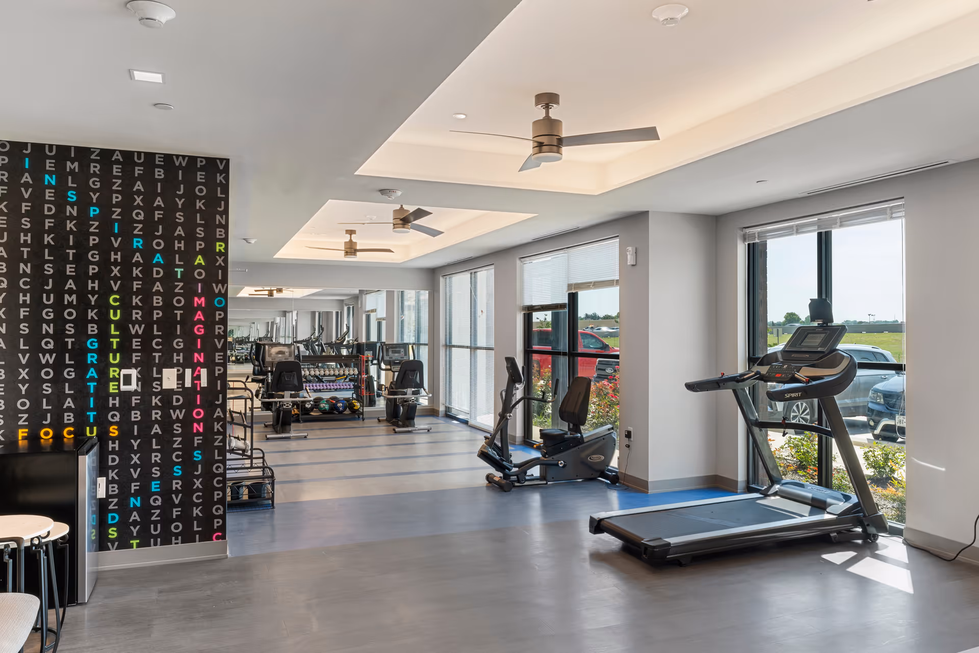A modern fitness room with exercise equipment including a treadmill, stationary bike, and weight rack. The room has large windows letting in natural light, ceiling fans, and a decorative wall featuring a colorful word search puzzle design.