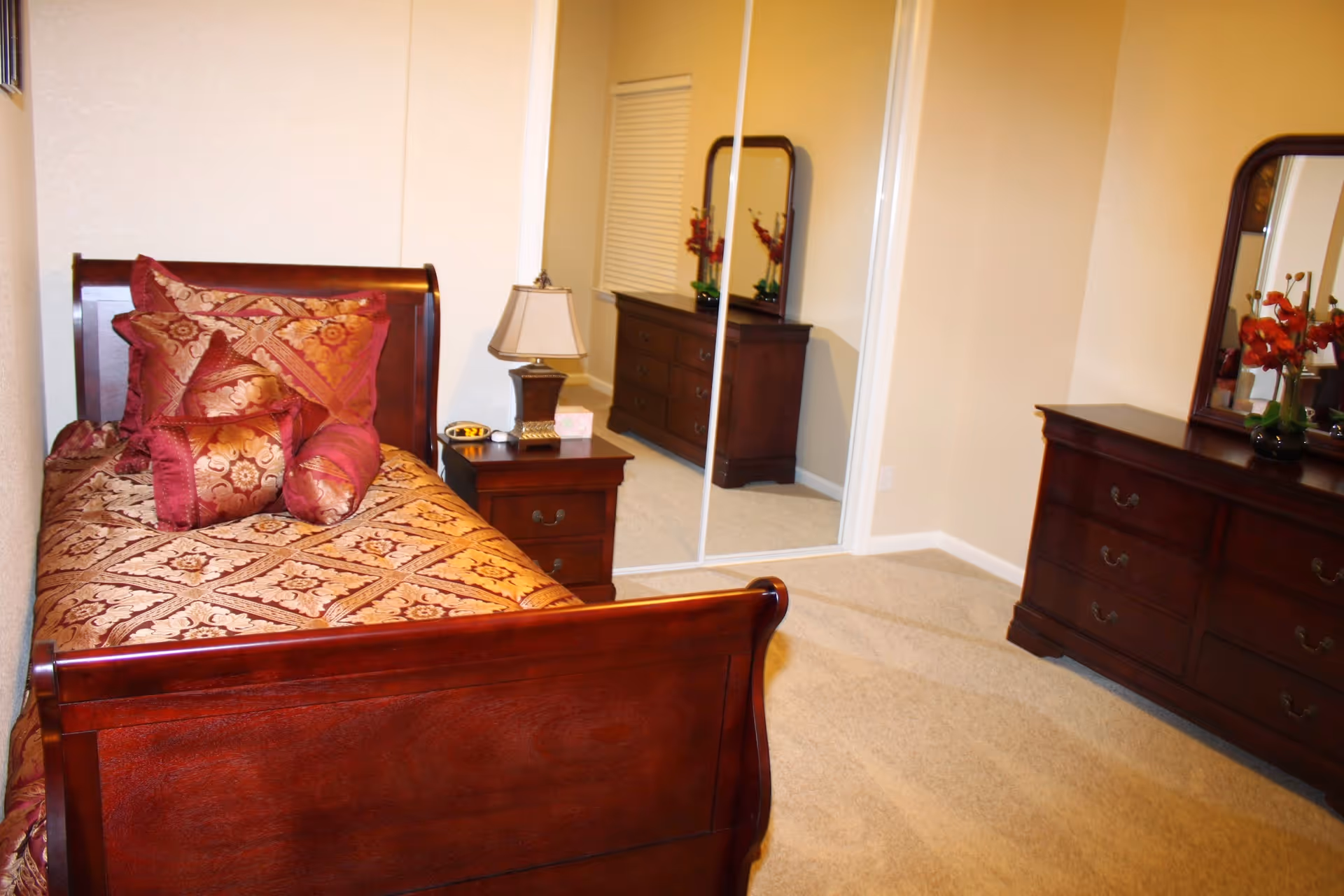 A bedroom with a wooden bed frame and a bed covered with a patterned bedspread and multiple decorative pillows. Next to the bed is a wooden nightstand with a lamp and a small dish. Across from the bed is a wooden dresser with a mirror and a vase with red flowers. There is a closet with sliding mirrored doors reflecting part of the room.