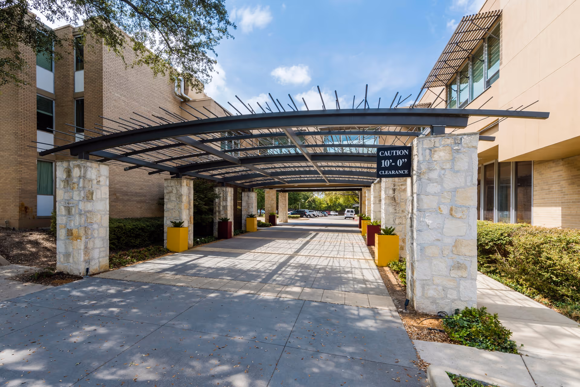 Outdoor covered driveway area with stone pillars and metal beams at a senior living facility named Autumn Leaves. The structure has a caution sign indicating 10 feet clearance. There are plants in yellow and maroon planters along the sides, with a building on either side and a parking lot visible in the background under a partly cloudy sky.